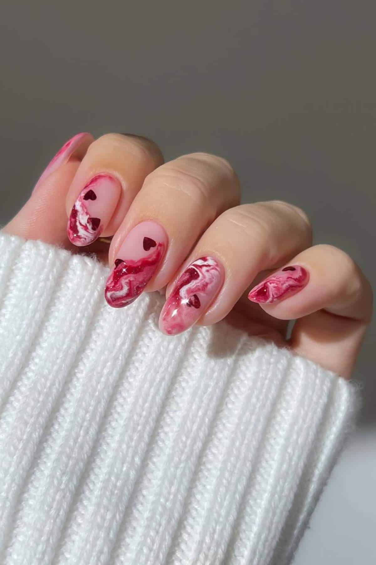A hand with long, almond-shaped nails featuring pink and red marble nail art and small heart designs, shown against a neutral background and a white ribbed sleeve.