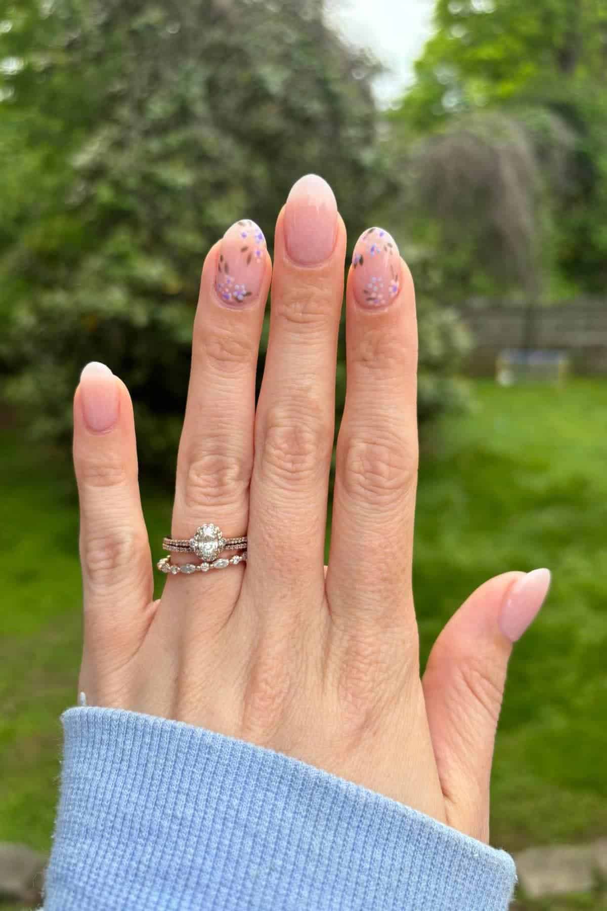 A hand with pink and purple floral nail art and three rings, including an engagement ring, is held up outdoors with a blurred green background.