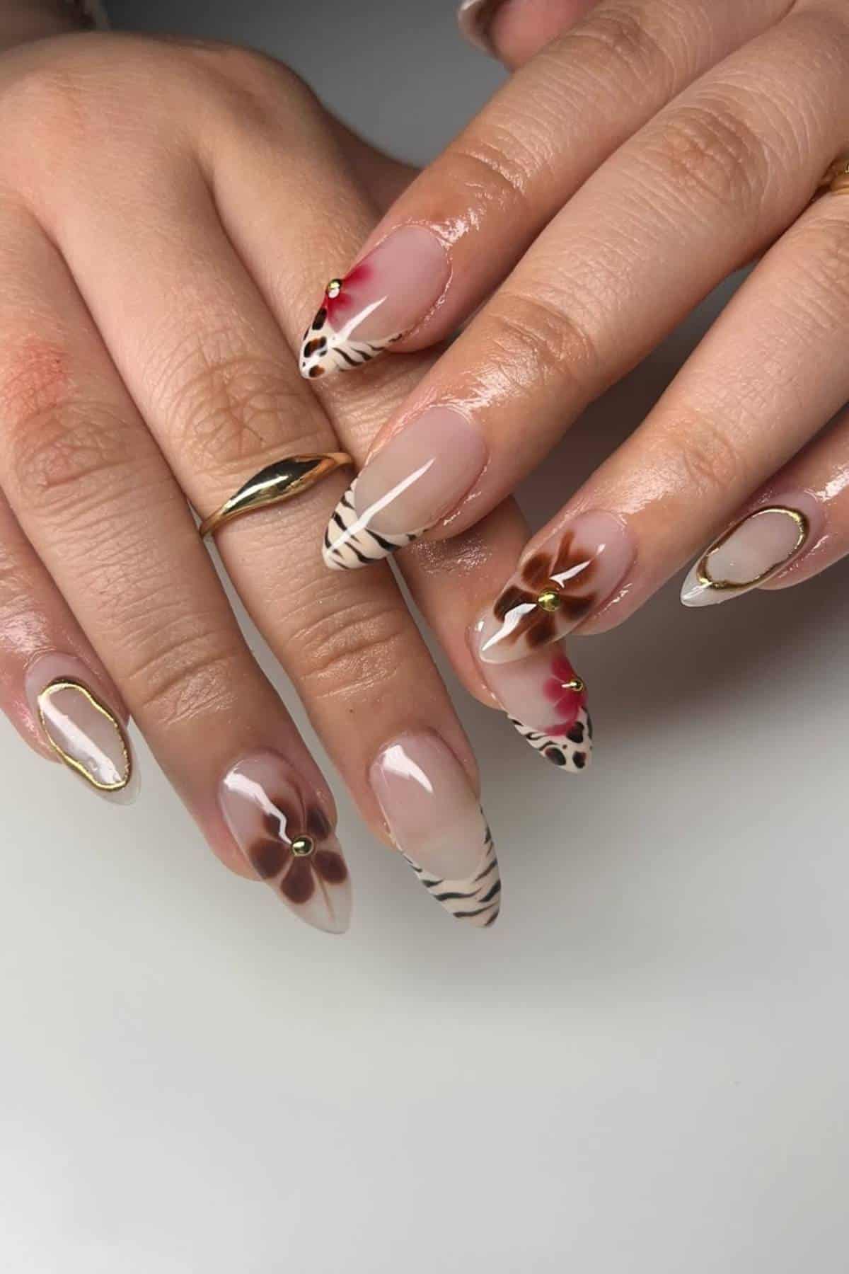 Hands with almond-shaped nails featuring floral designs, gold accents, red gems, and animal print patterns on a neutral base.