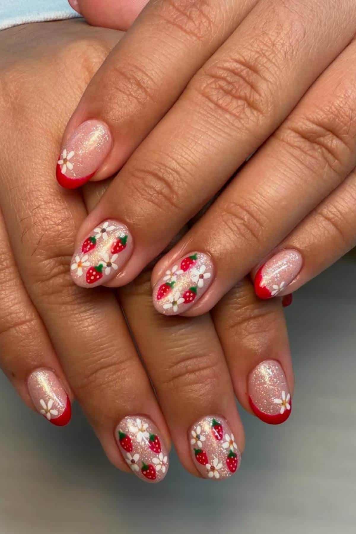 Hands with short, almond-shaped nails featuring a shimmery nude base, red French tips, and nail art of strawberries and white flowers.
