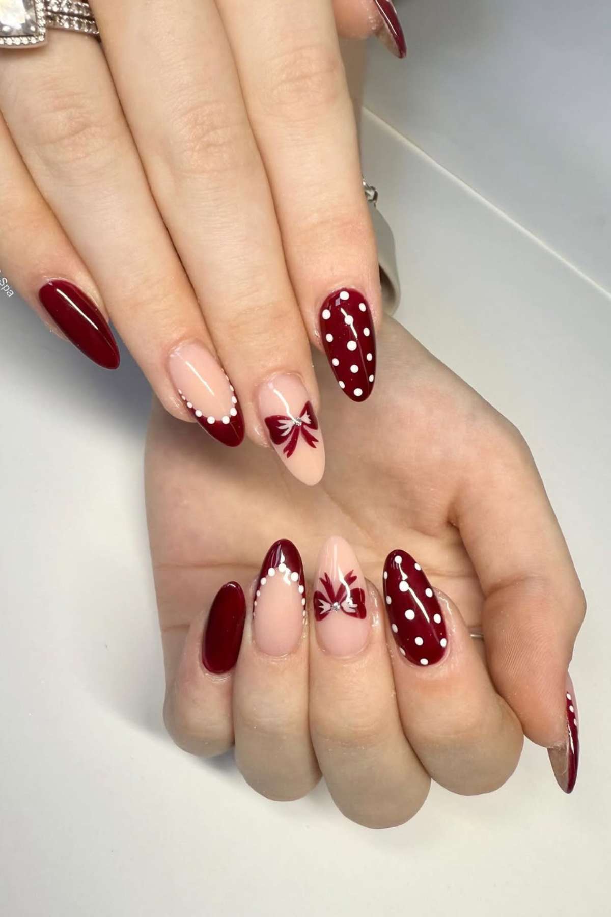 Hands with almond-shaped nails painted in burgundy and nude, featuring white polka dots and red bow designs as nail art.