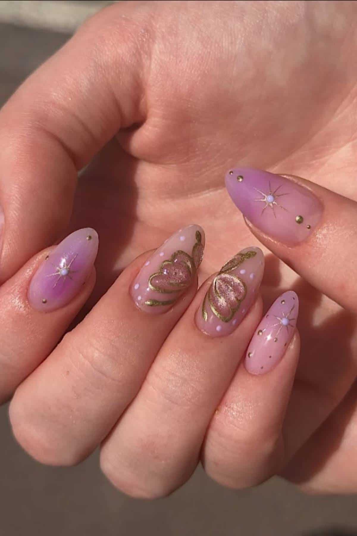 A hand with almond-shaped nails painted in pink and purple shades, decorated with gold hearts, stars, and small dots.