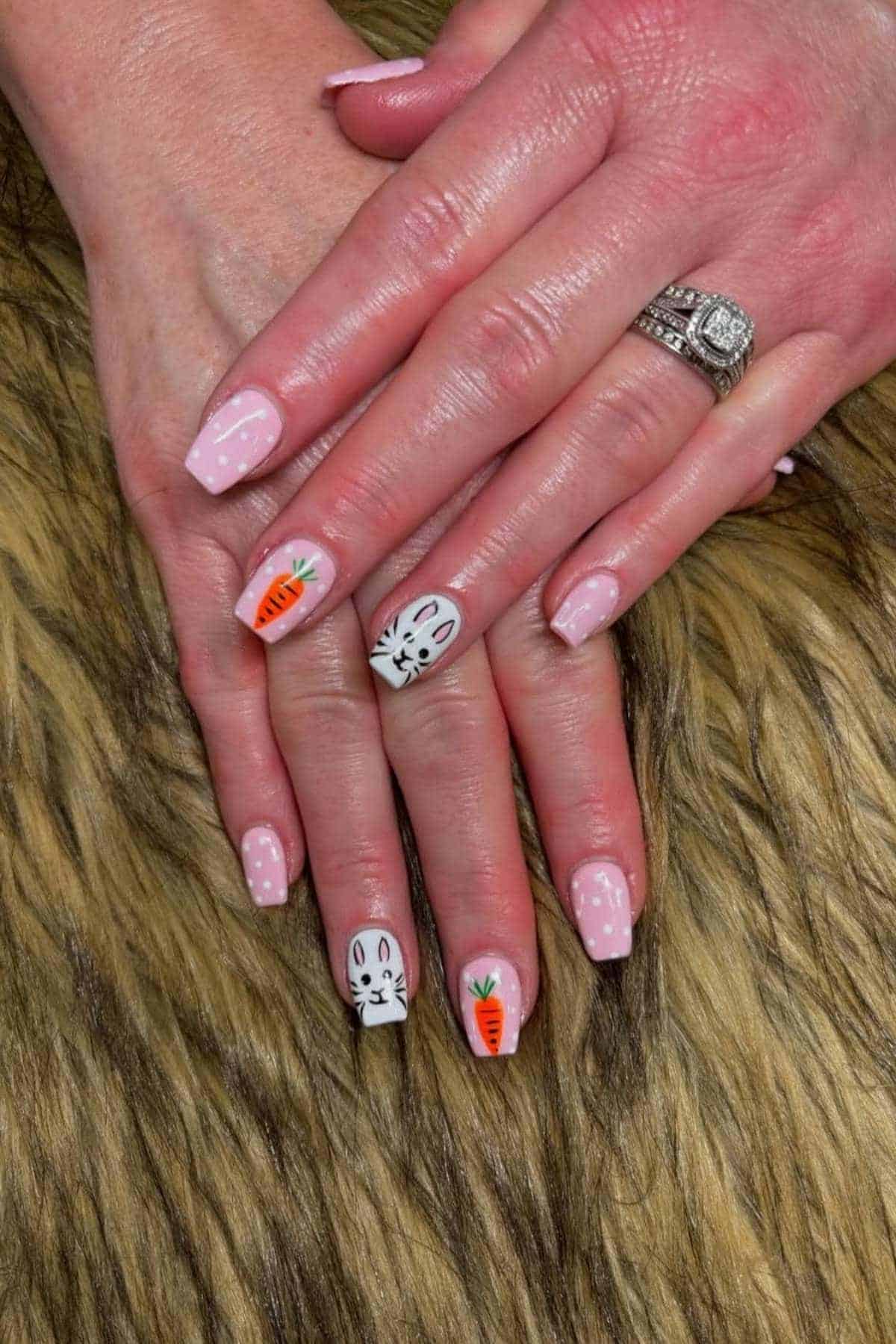Hands with light pink manicured nails; two nails feature white bunny faces and two have orange carrots drawn on them, displayed on a furry brown surface.