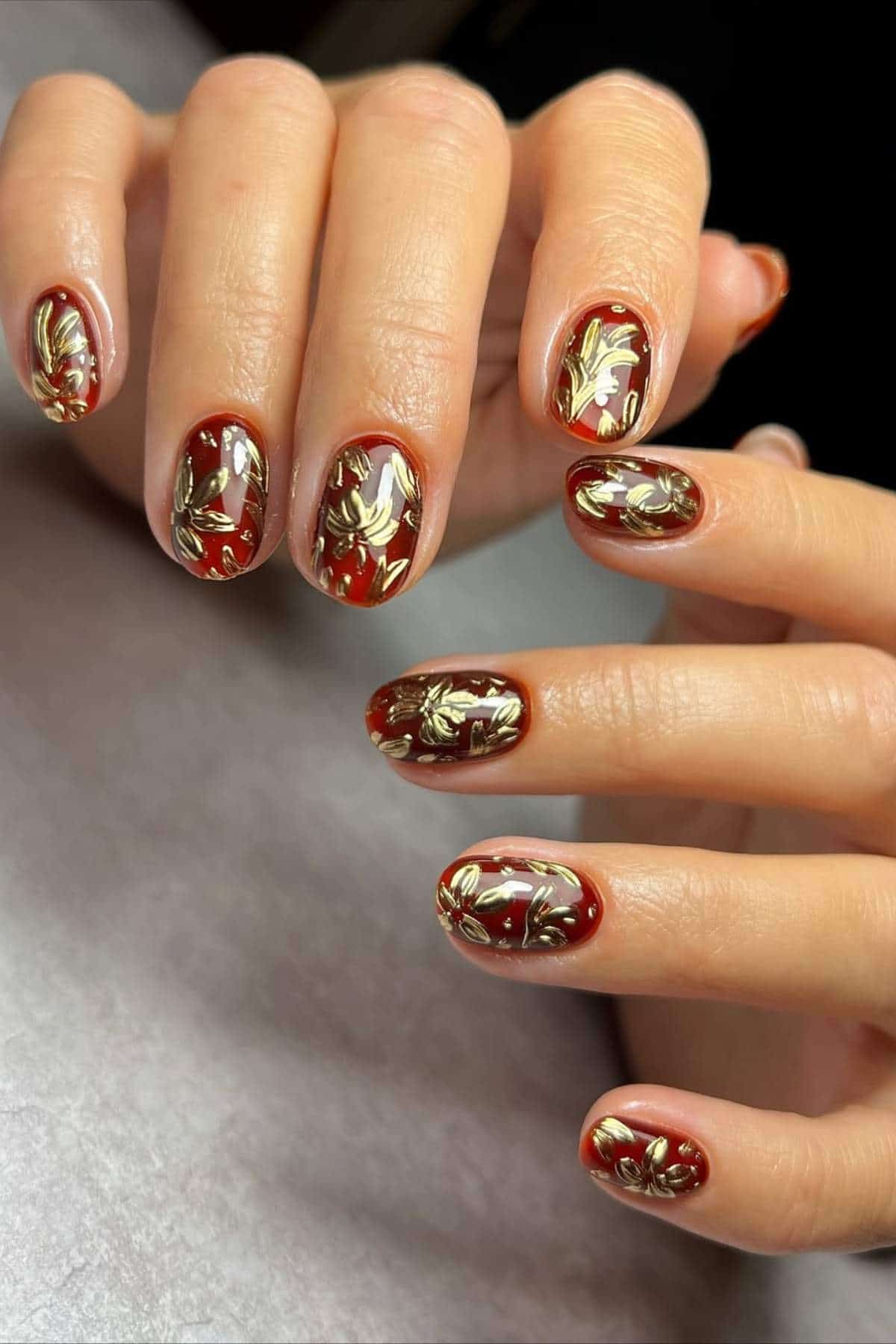 Hands with short oval nails painted dark red and decorated with gold leaf floral designs, displayed on a light gray surface.