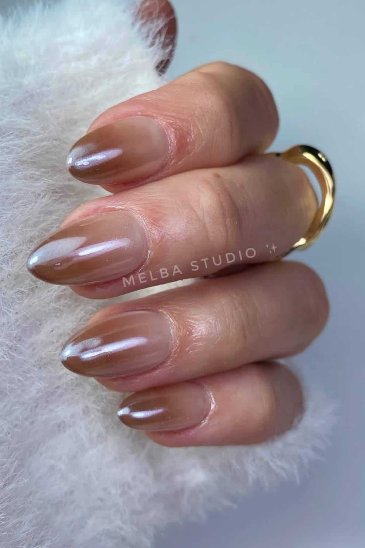A hand with long, almond-shaped nails featuring a glossy, light brown and nude ombre polish, resting on a white fluffy surface.