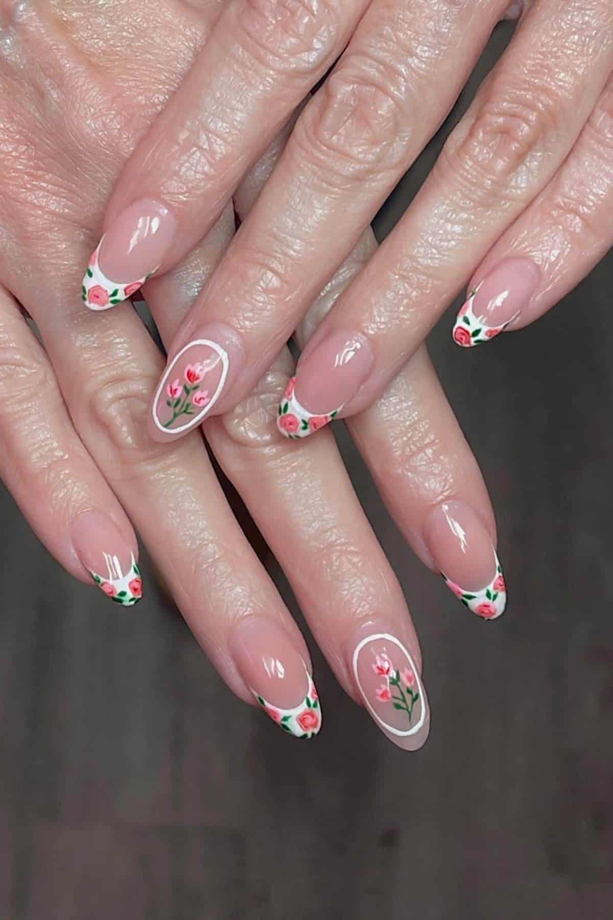 Hands with almond-shaped nails featuring a French manicure with small red and green floral designs on the tips and an accent nail with a full flower on a light pink base.
