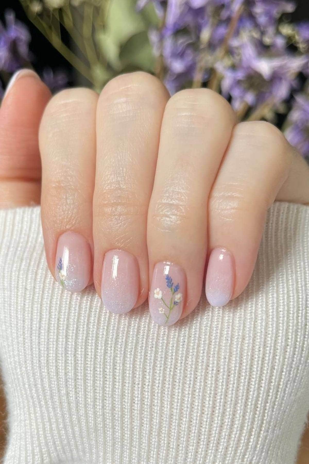 A hand with short, neatly manicured nails painted in a pale pink ombre with small floral designs on two nails. The person is wearing a light-colored ribbed sleeve.