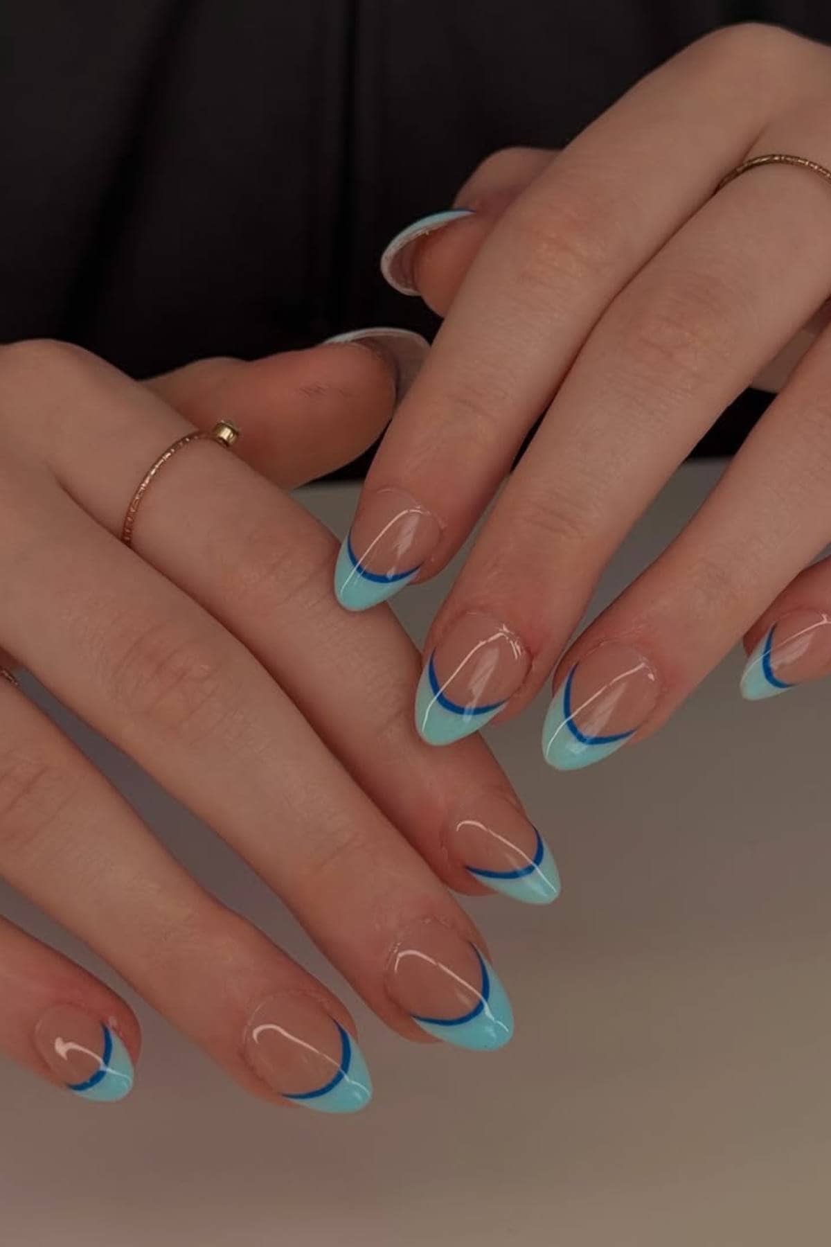 Hands with almond-shaped nails featuring a clear base and light blue tips outlined with a thin, dark blue line. The person is wearing thin gold rings.