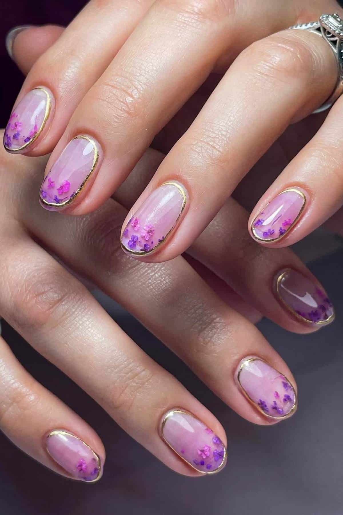 Close-up of hands with oval-shaped nails featuring a pink base, purple floral designs, and gold outlines near the cuticles.