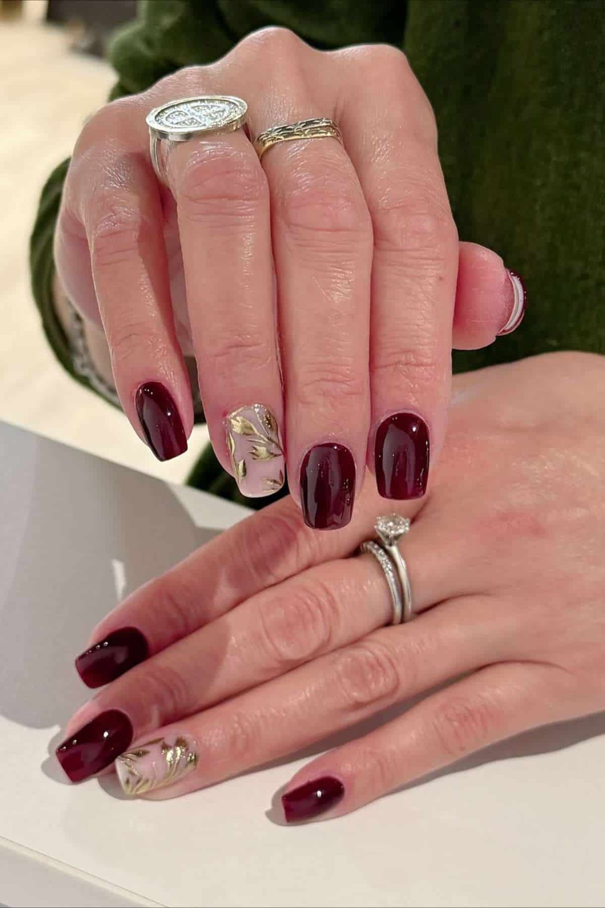 Hands with dark red manicured nails; accent nails have gold leaf designs. The person is wearing silver rings on both hands and a green long sleeve top.