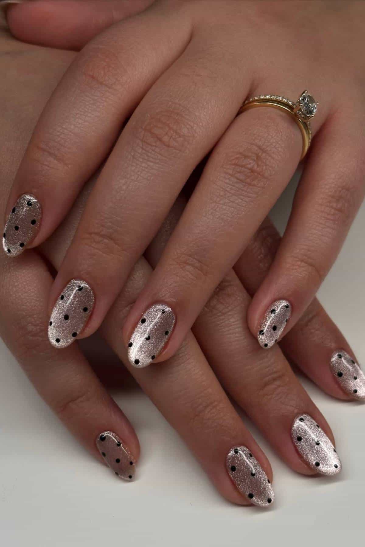Close-up of hands with short, almond-shaped nails painted in a shimmery nude polish and decorated with black polka dots; one hand wears a gold ring with a single clear stone.