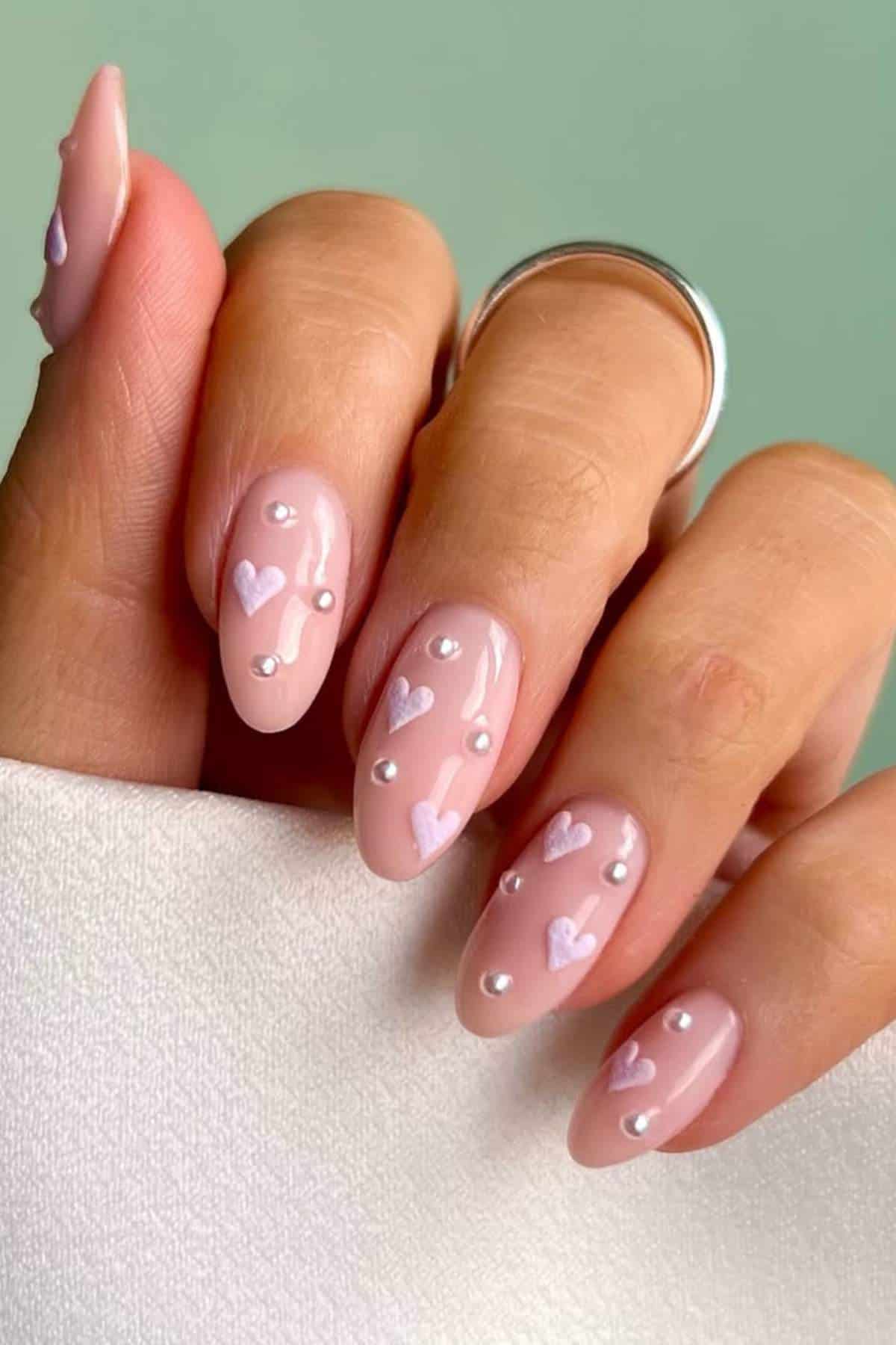 A hand with almond-shaped nails painted in a nude pink color with small white hearts and pearl-like bead accents.