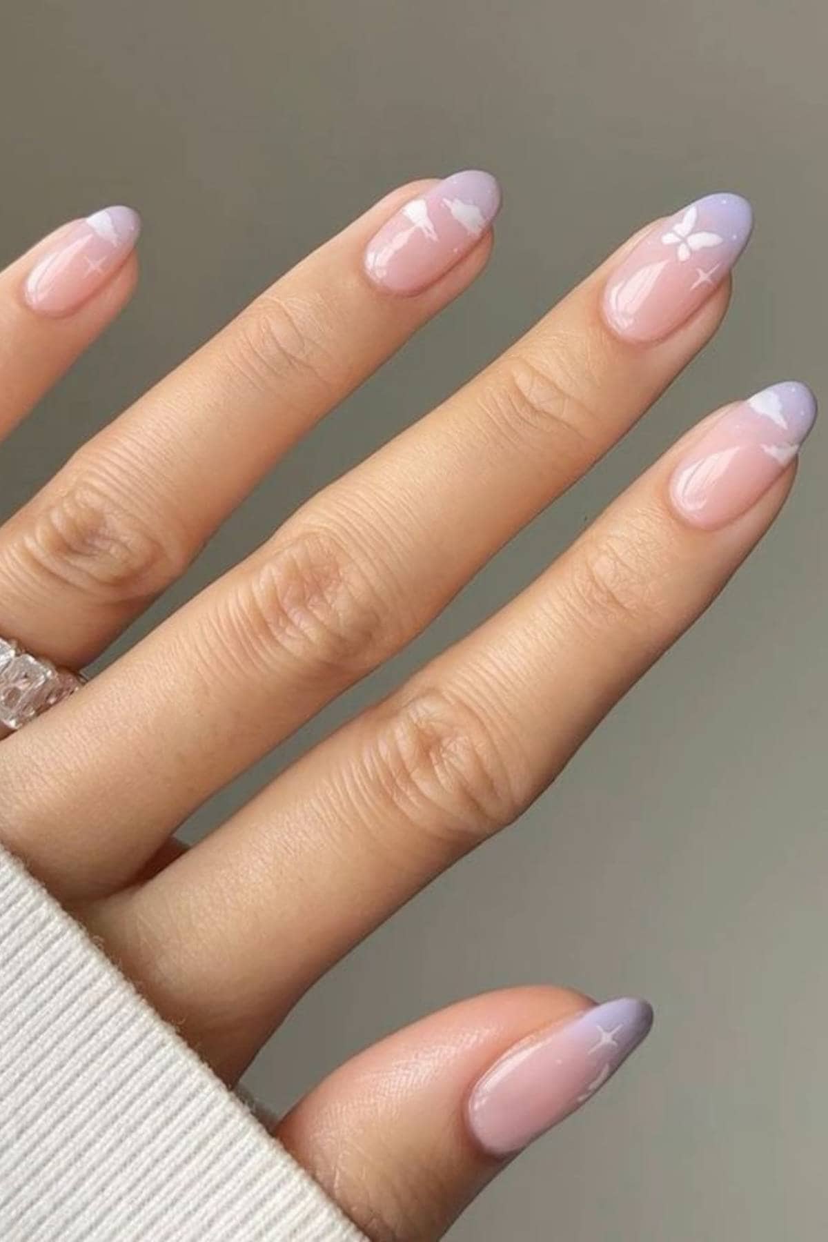 A hand with almond-shaped nails featuring a glossy nude base, purple tips, and white cross designs on two nails. The person wears a clear ring and a light-colored sleeve.