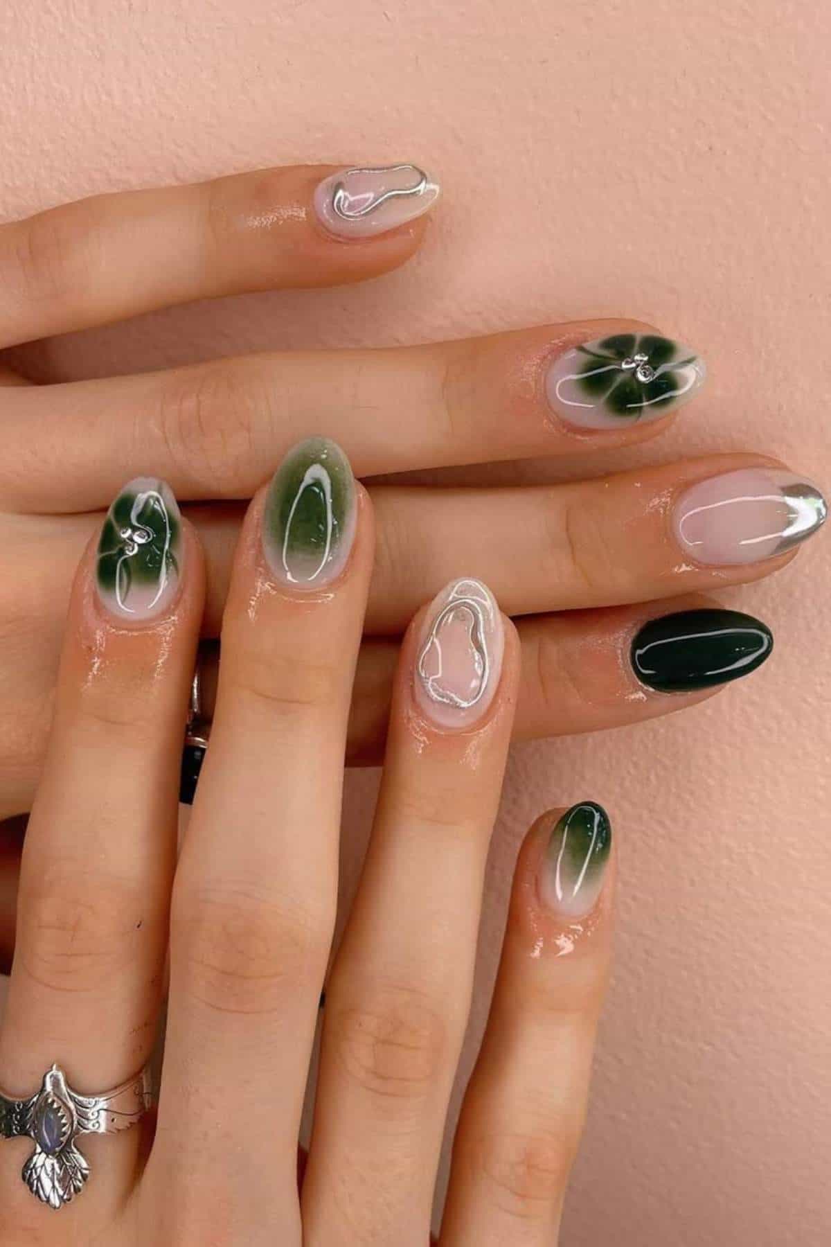 A hand with almond-shaped nails featuring green, white, and clear abstract designs, including marble and leaf patterns, against a light pink background.