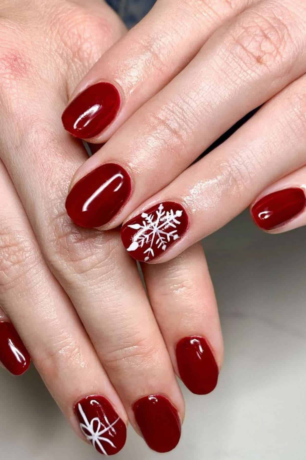 Hands with short, glossy red nails; two nails feature white snowflake designs as accent nail art.