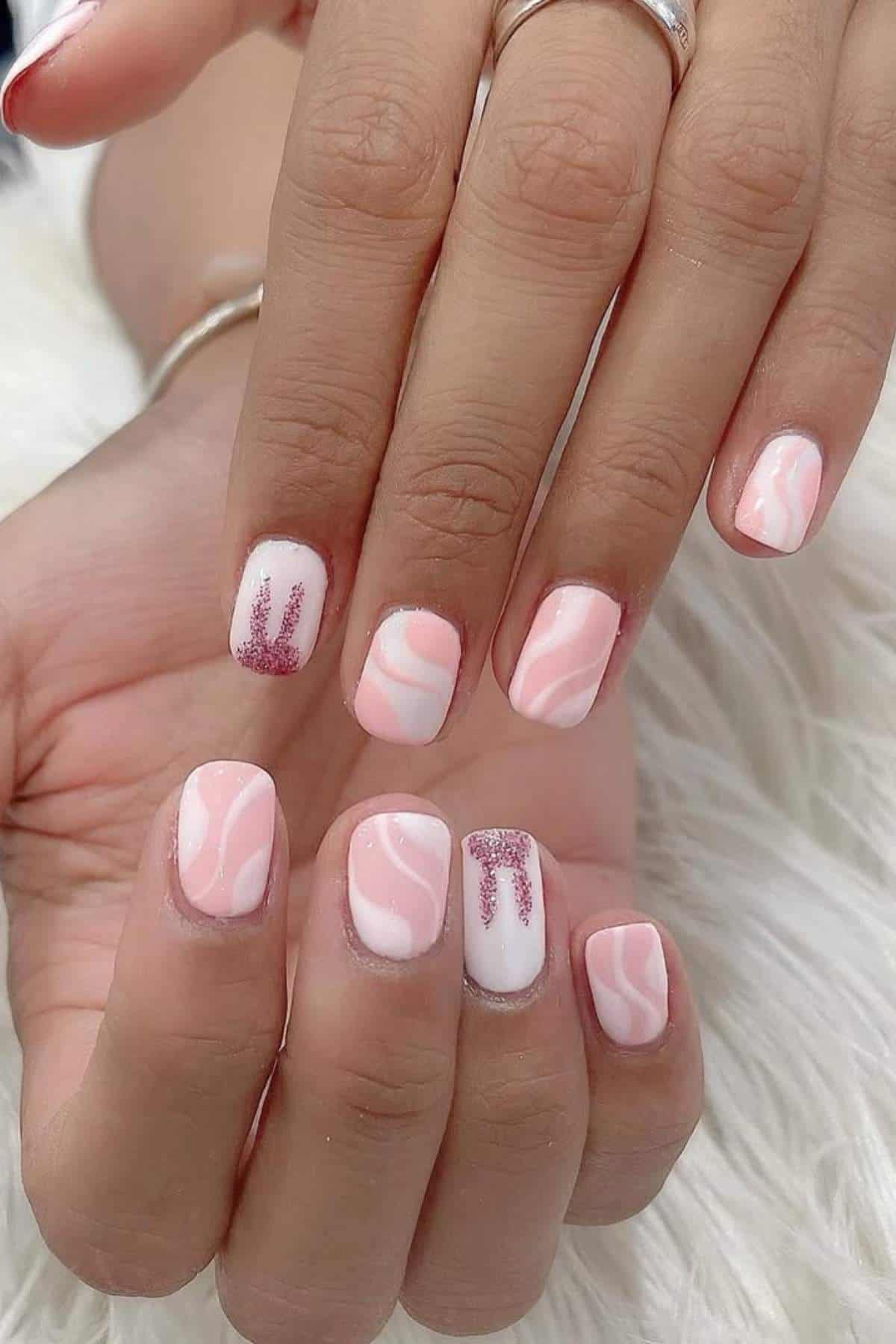 Hands with short square nails featuring a pink and white abstract swirl design, with two nails accented by pink glitter and pi symbol art.