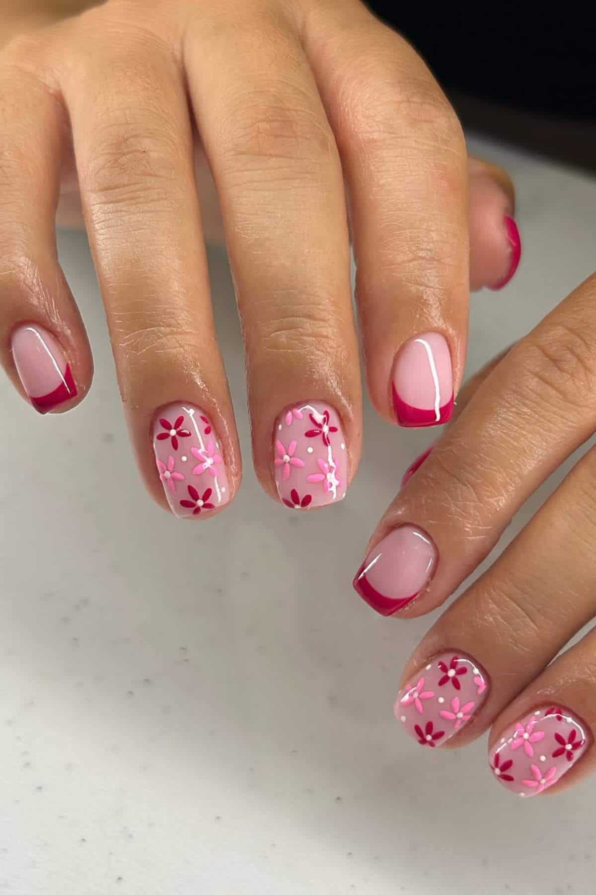 Hands with manicured nails featuring pink polish, red tips, and alternating floral designs with red flowers on a light pink background.