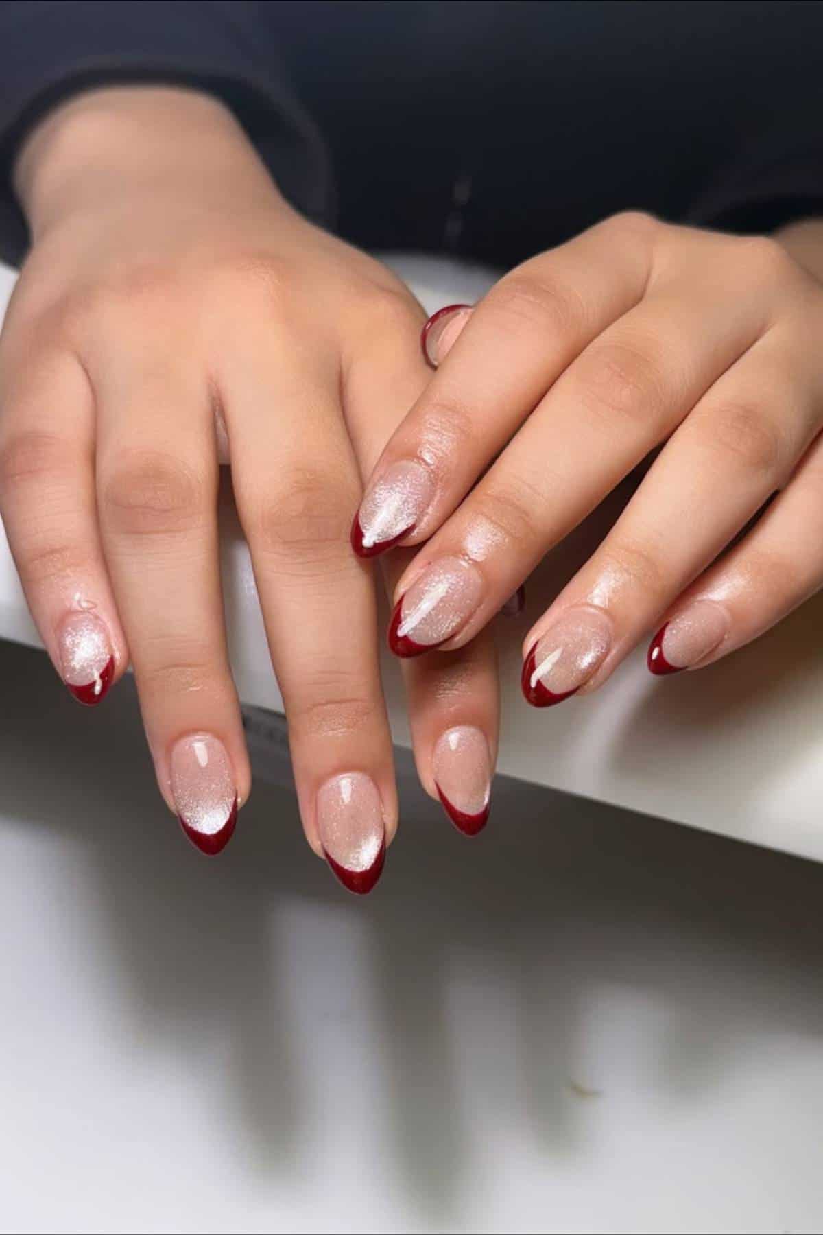 Two hands with almond-shaped nails featuring a nude base and metallic shimmer, each nail tipped with a deep red color.