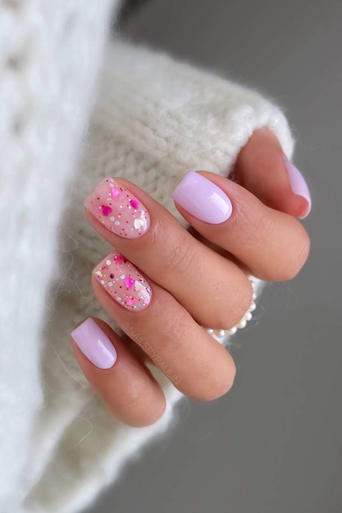 A hand with short, rounded nails painted light lavender; two nails feature pink and white glitter and confetti accents.