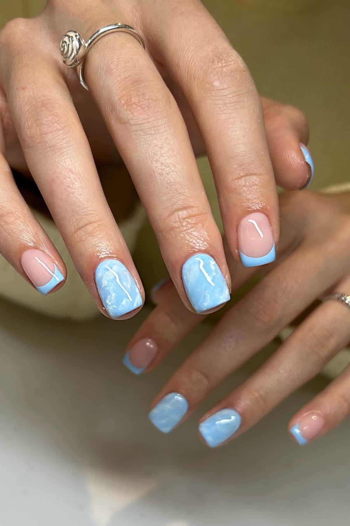 Hands showing a manicure with short square nails; some nails have a blue marble design, others have a pale pink base with blue tips.