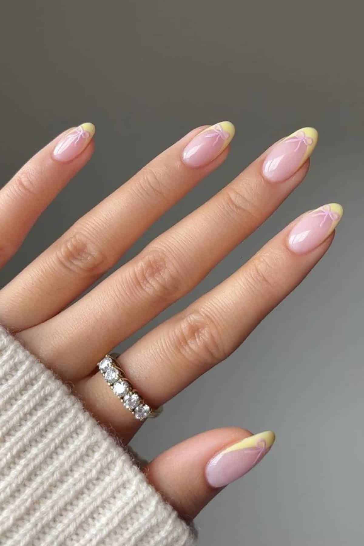 A hand with almond-shaped nails featuring a light pink base and yellow tips with subtle line art, wearing a diamond ring and a beige sweater.