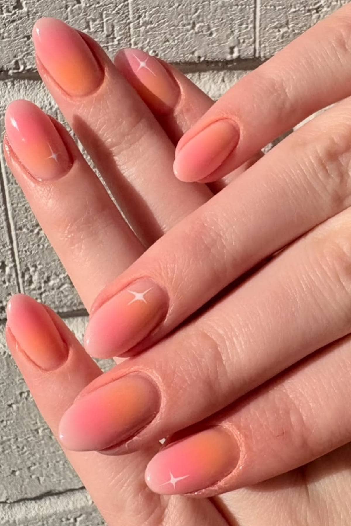 A hand with almond-shaped nails painted in a pink and orange gradient with subtle white star accents, shown against a textured light-colored wall.