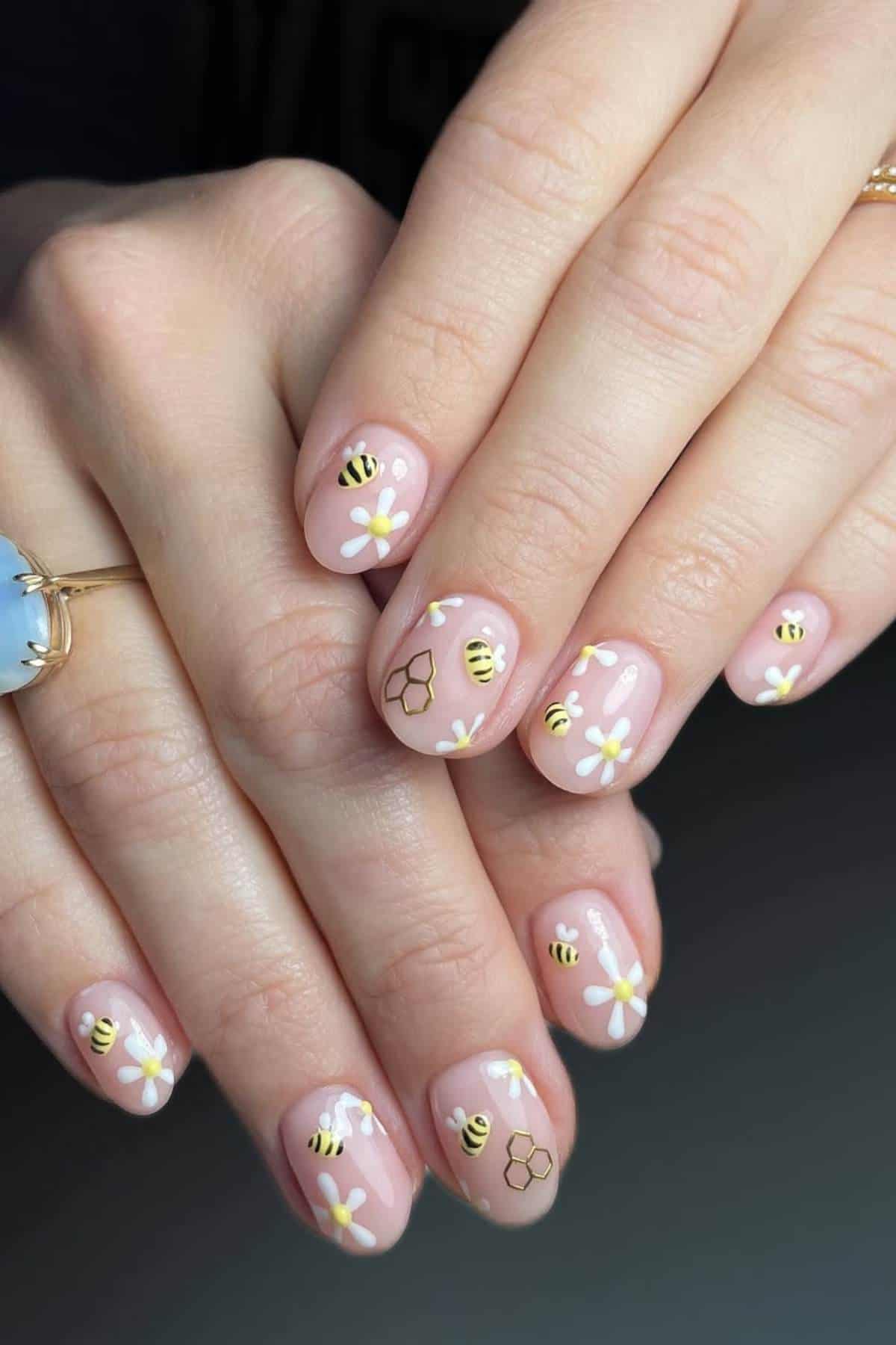 Close-up of hands with light pink nail polish featuring painted bees, white flowers, and honeycomb designs on short, rounded nails.