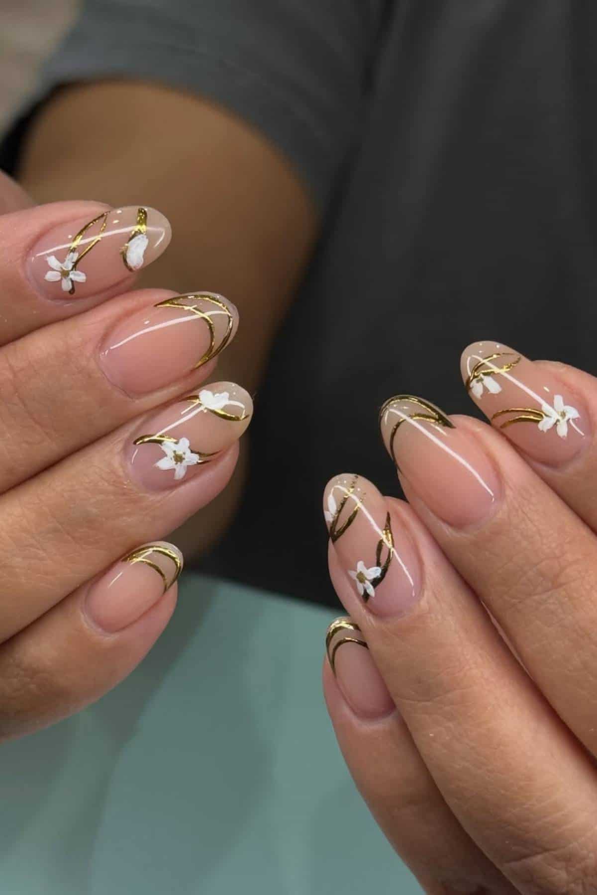 Hands with almond-shaped nails featuring clear polish, gold lines, and small white flower designs on each nail.