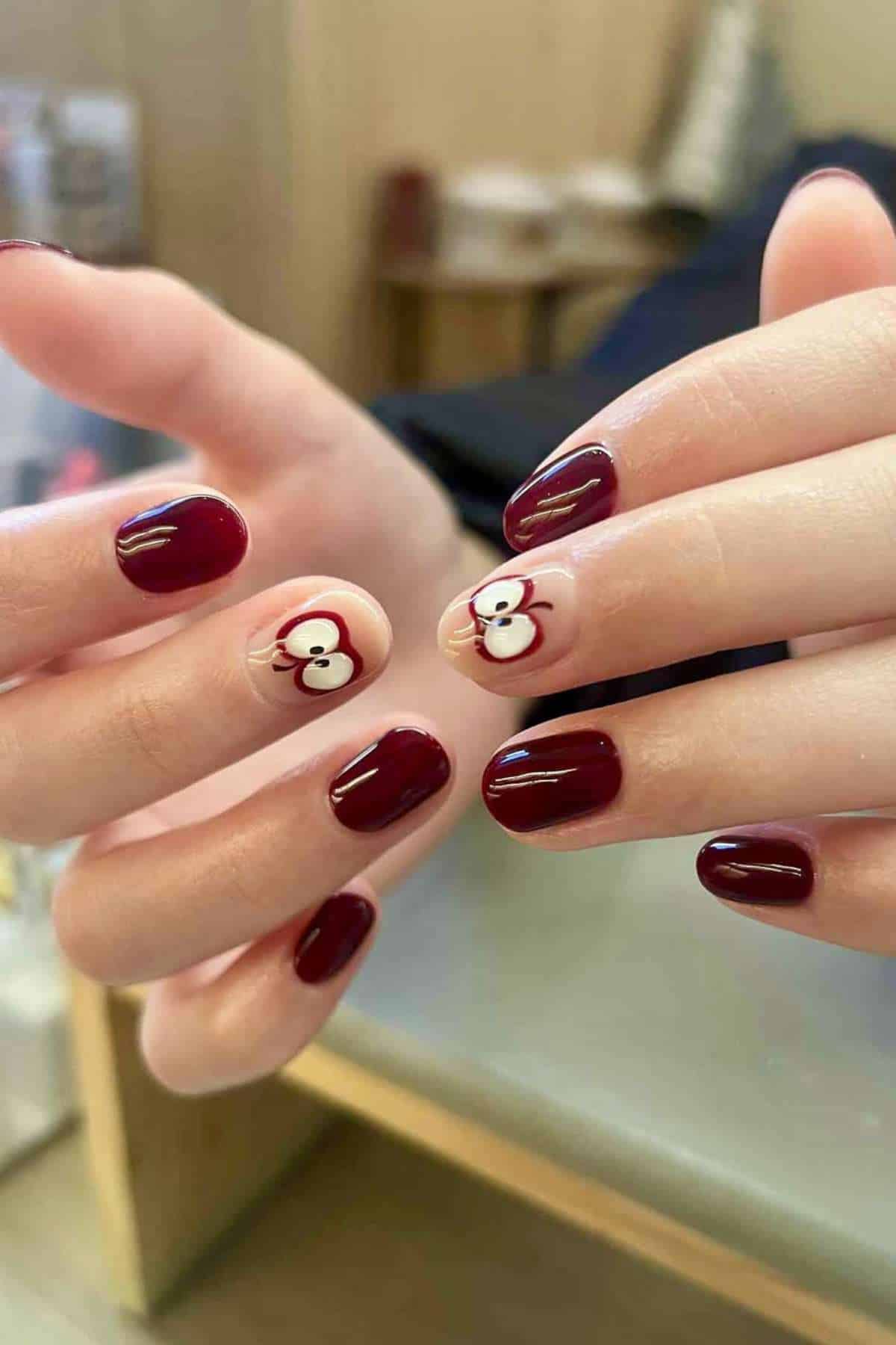 Hands with short, rounded nails painted dark red; thumbs feature nail art of a simple white and red cartoon mouse face with black whiskers.