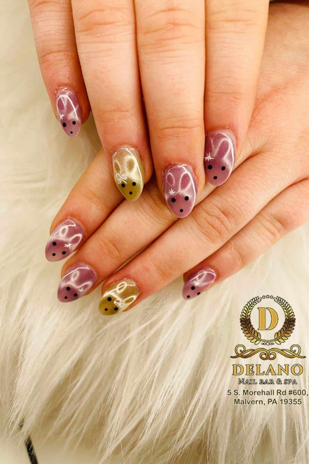 Close-up of hands with almond-shaped nails painted in pink and gold, featuring bunny face designs with black dots for eyes and white accents. Delano Nail Bar & Spa logo is visible.
