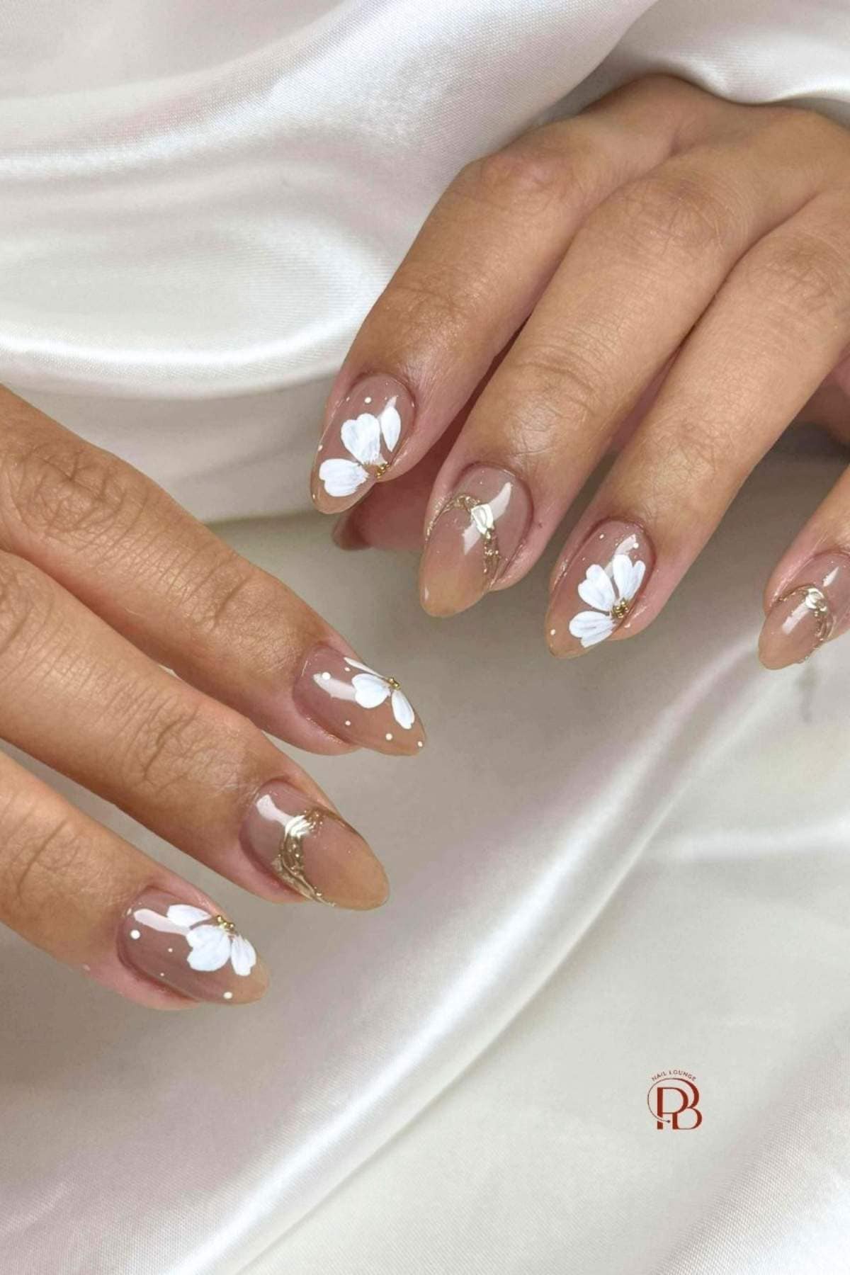 Hands with almond-shaped nails featuring a nude base, white floral designs, gold accents, and small white dots, resting on a white satin fabric.