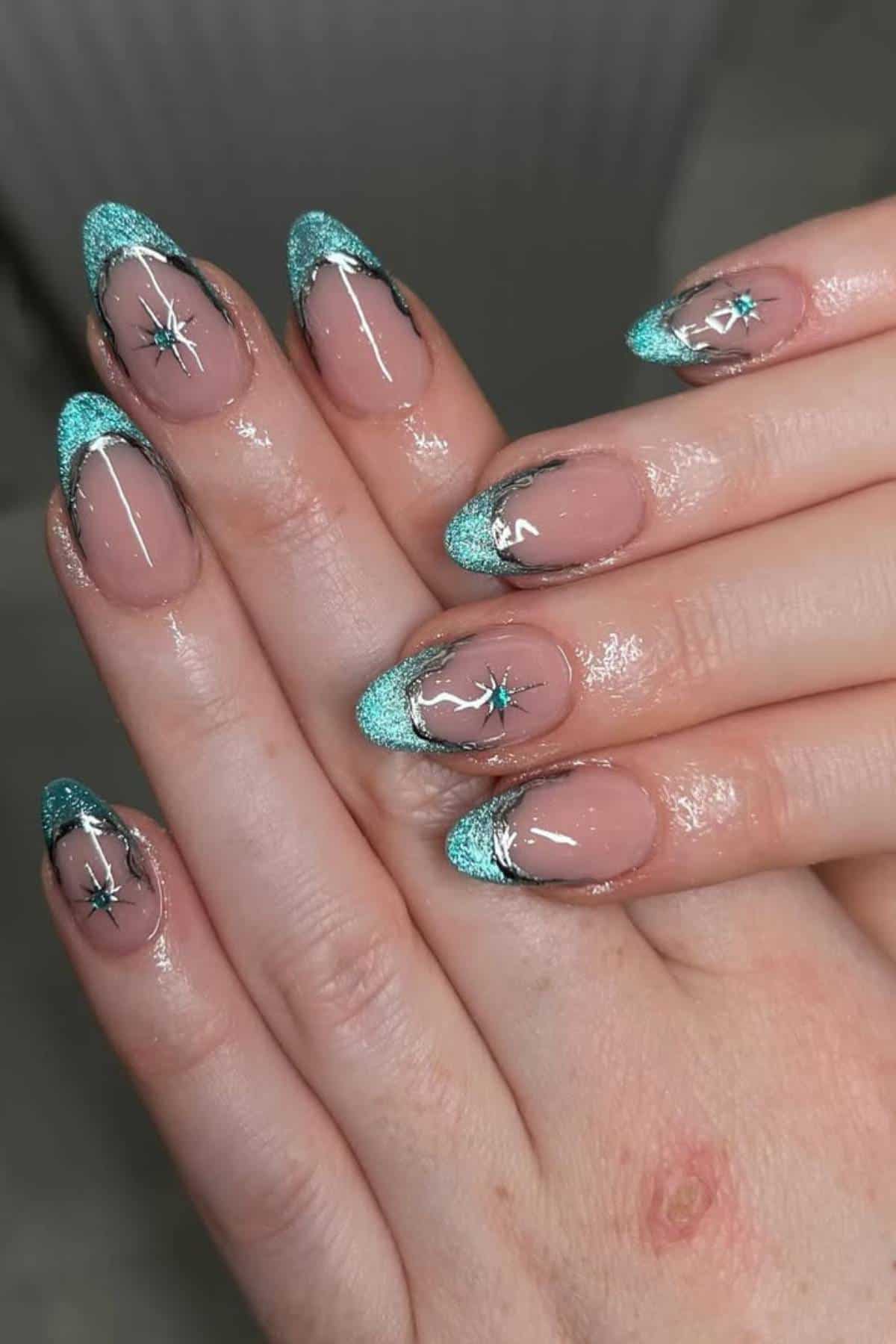 Hands with almond-shaped nails featuring turquoise glitter French tips, black and silver lines, and starburst nail art designs on a nude base.