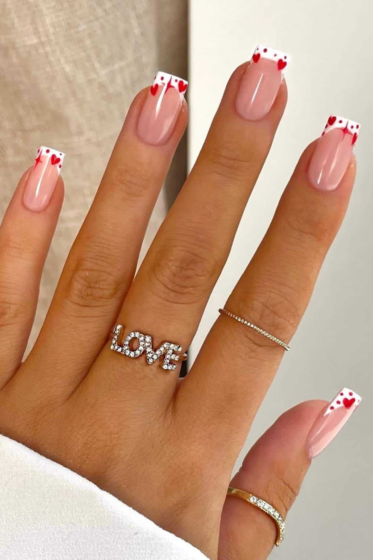 A hand with nude-colored nails featuring white French tips decorated with small red heart designs; the person wears rings, including one that spells "LOVE.