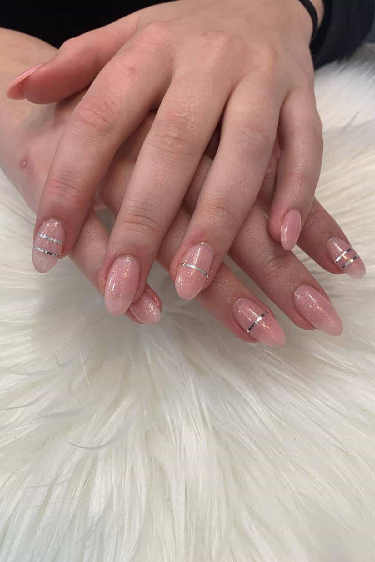 Hands with almond-shaped nails painted in light pink polish, featuring subtle glitter and thin silver horizontal stripes on some nails, resting on a white textured surface.