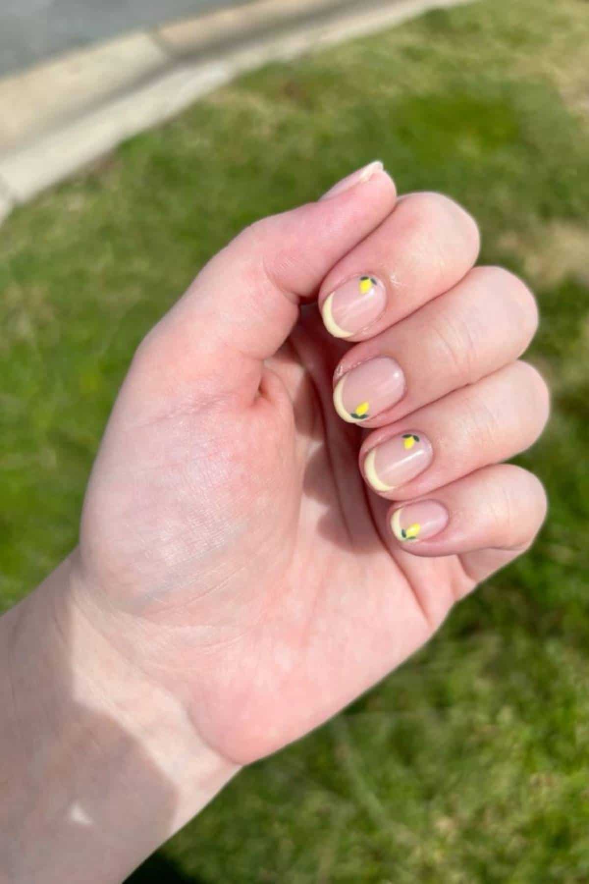 A hand with manicured nails featuring a nude base, yellow tips, and small yellow and black flower designs, shown outdoors over green grass.