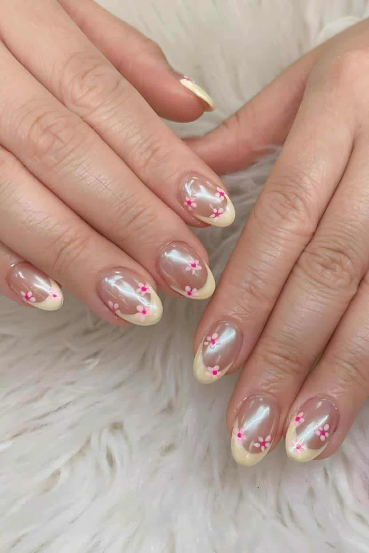 Hands with almond-shaped nails featuring a pearly nude base, pale yellow French tips, and small pink and white floral designs, resting on a white textured surface.
