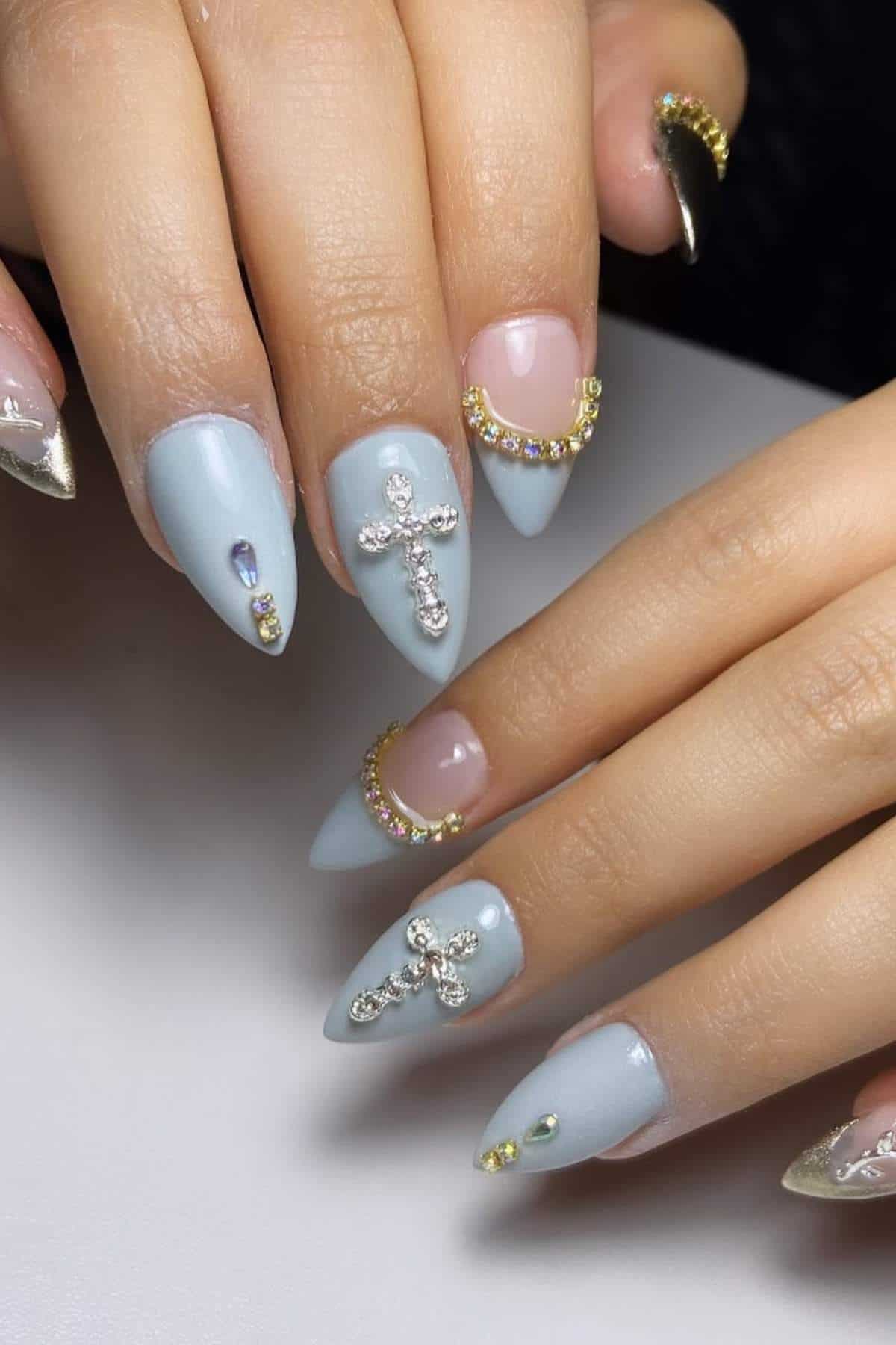 Hands with long, pointed, light blue nails featuring rhinestone crosses, gold and rhinestone accents, and metallic gold tips.