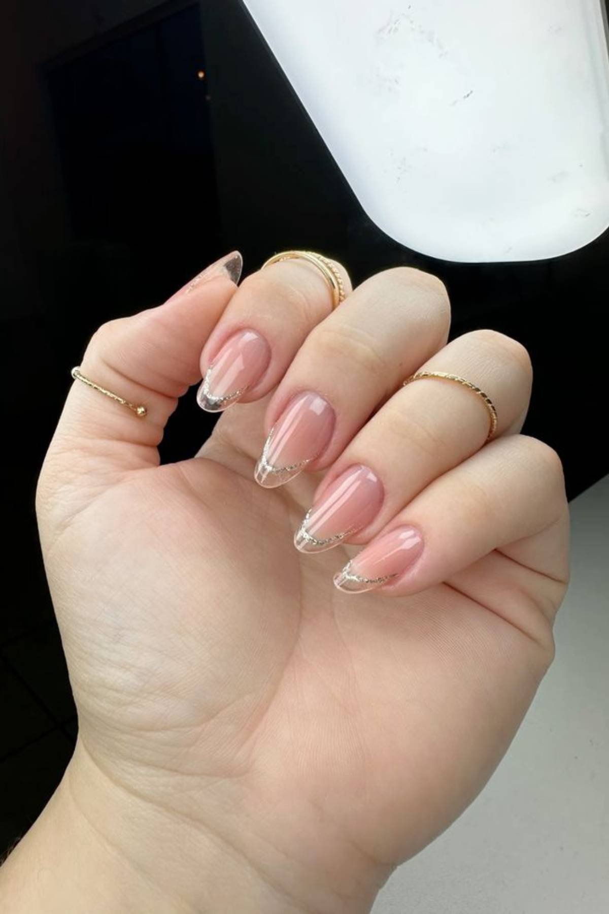 A hand with long almond-shaped nails featuring a nude pink base and thin, clear tips, adorned with gold rings on multiple fingers.