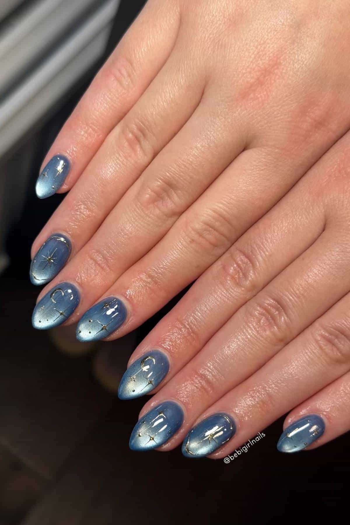 A hand with medium-length, almond-shaped nails painted in blue with gold star and constellation designs.
