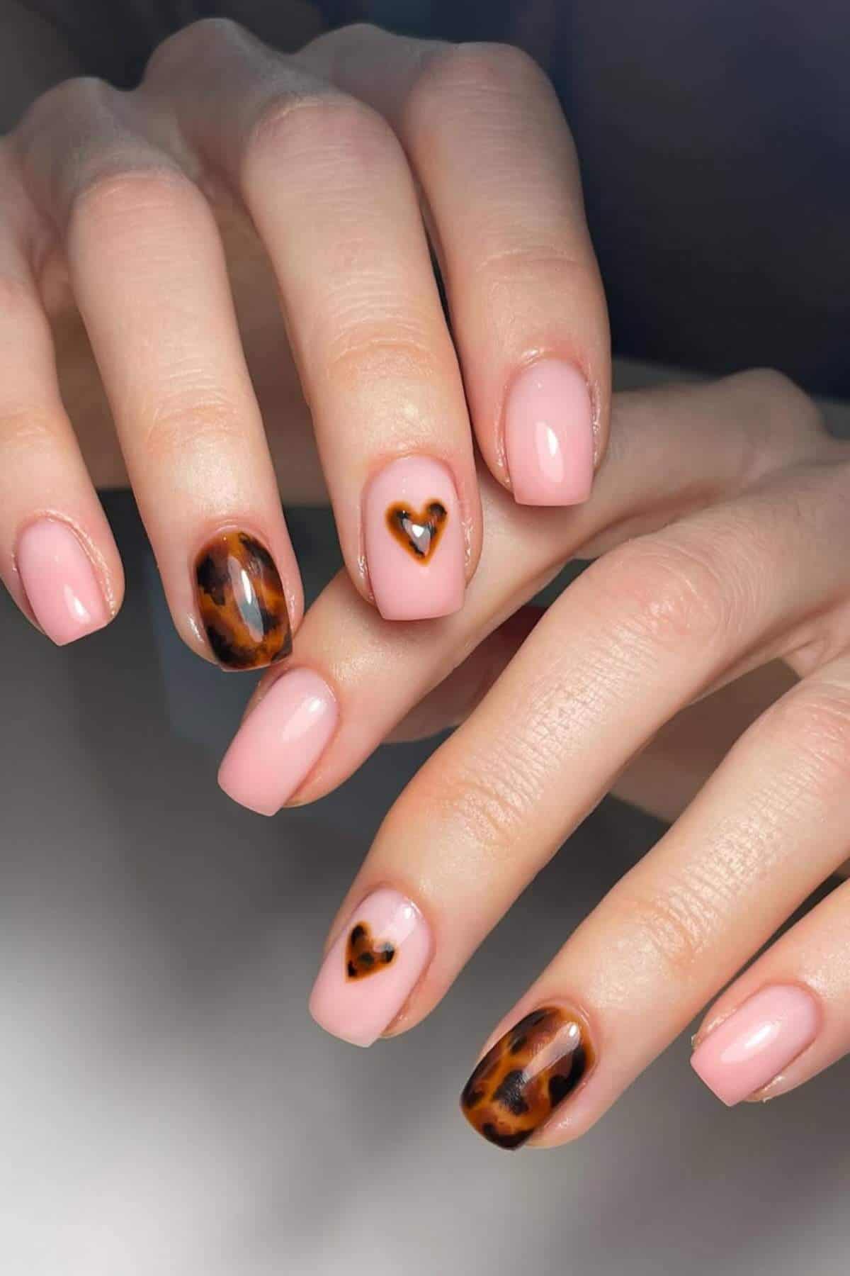 Hands with manicured nails; most nails are painted light pink, with two featuring tortoiseshell patterns and heart shapes on the ring and middle fingers.
