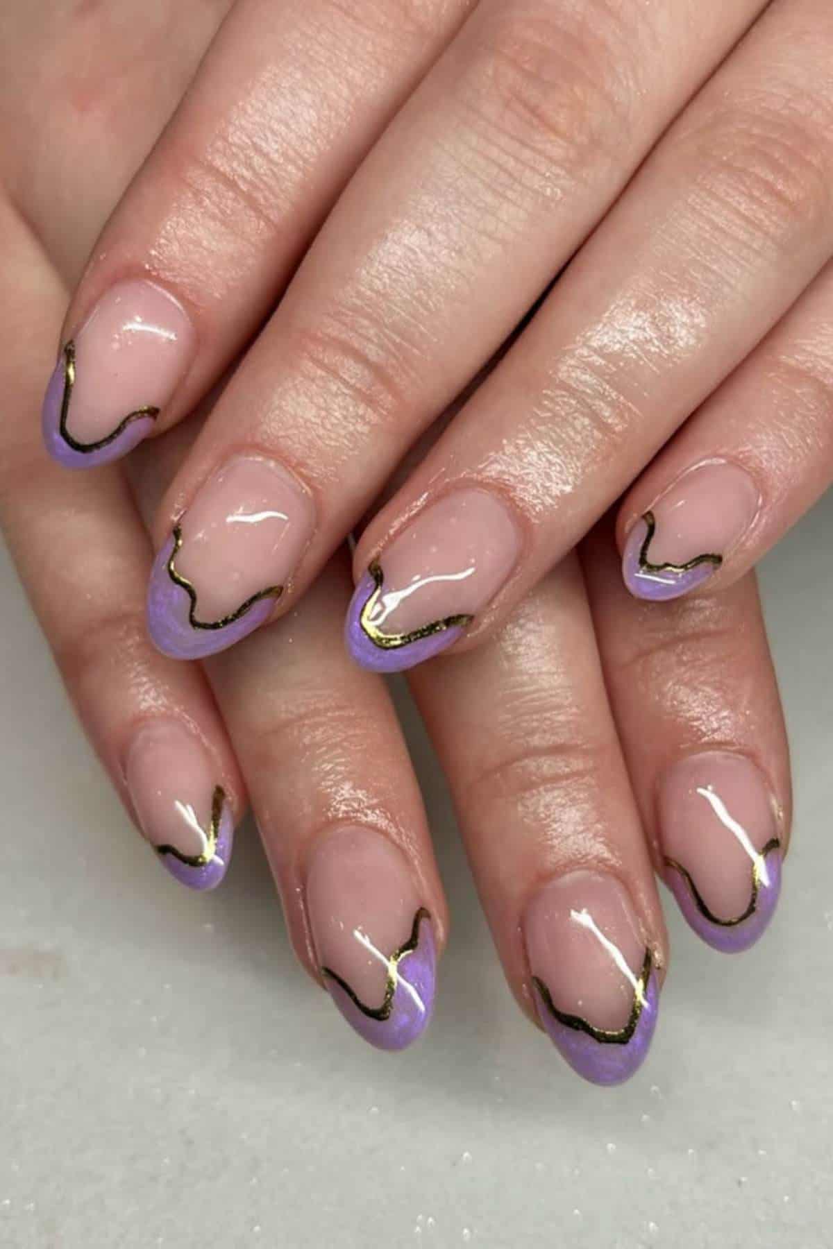 Hands with short, almond-shaped nails featuring a nude base, purple wavy tips, and irregular gold outlines as nail art design.