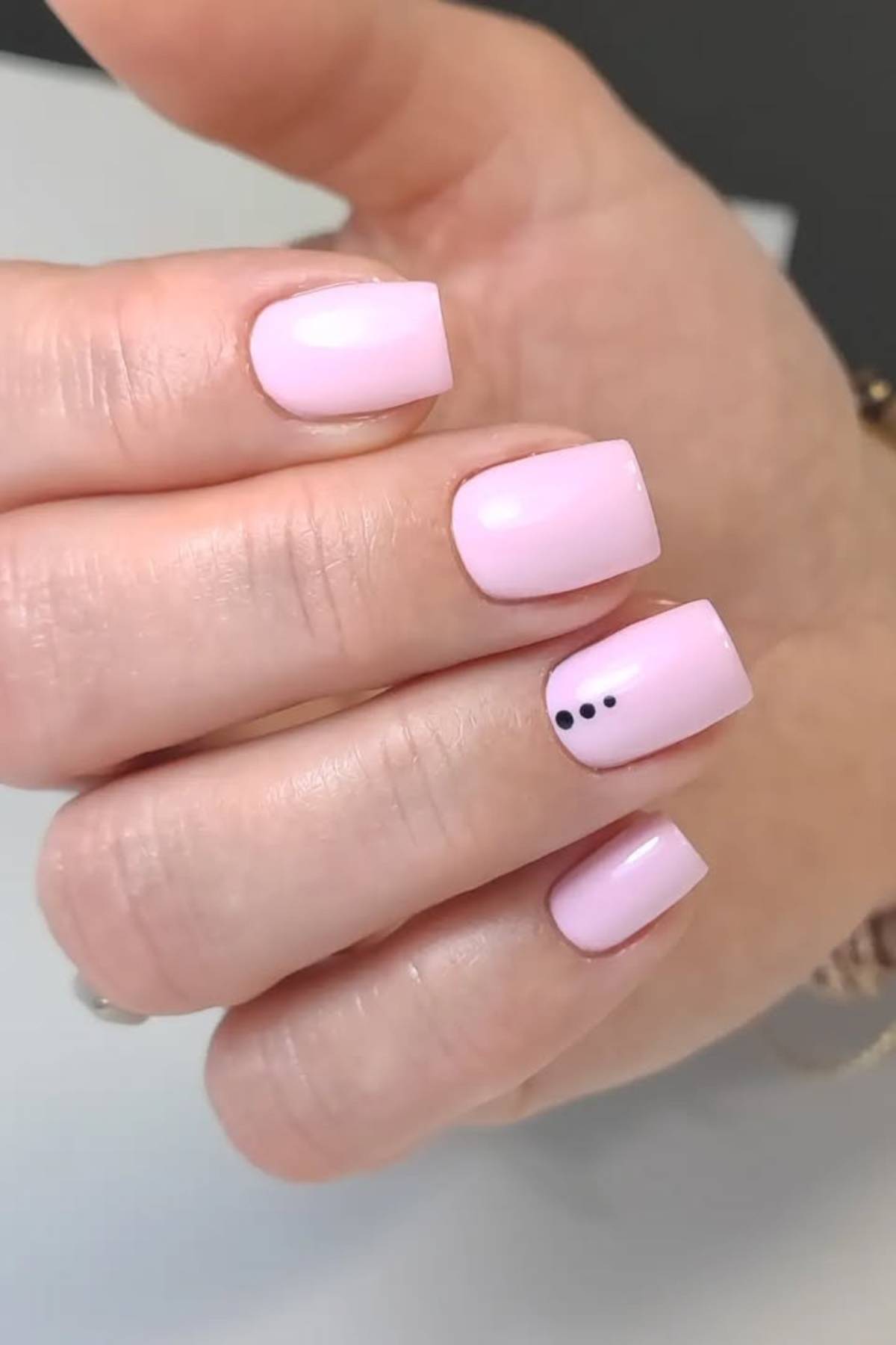A hand with short, square-shaped nails painted light pink; the ring fingernail features three small black dots near the tip.