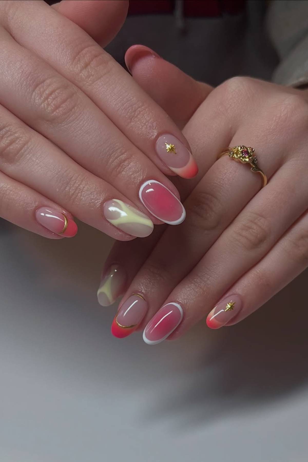 Hands with almond-shaped nails featuring pink, white, yellow, and red designs, gold lines, and star accents. The person wears a gold ring with a decorative detail.