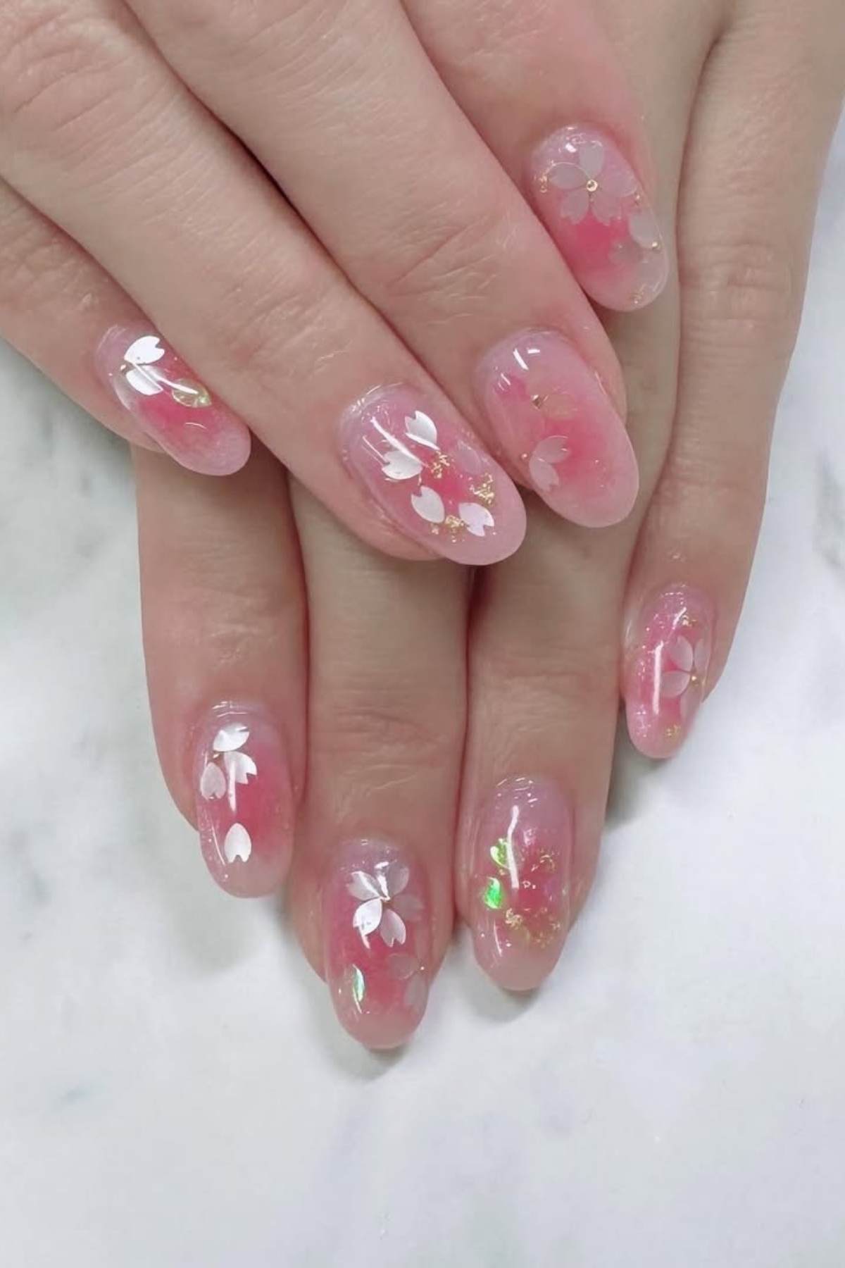 Hands with oval-shaped nails featuring pink translucent polish and white floral designs, accented with small gold and iridescent details, resting on a white marble surface.