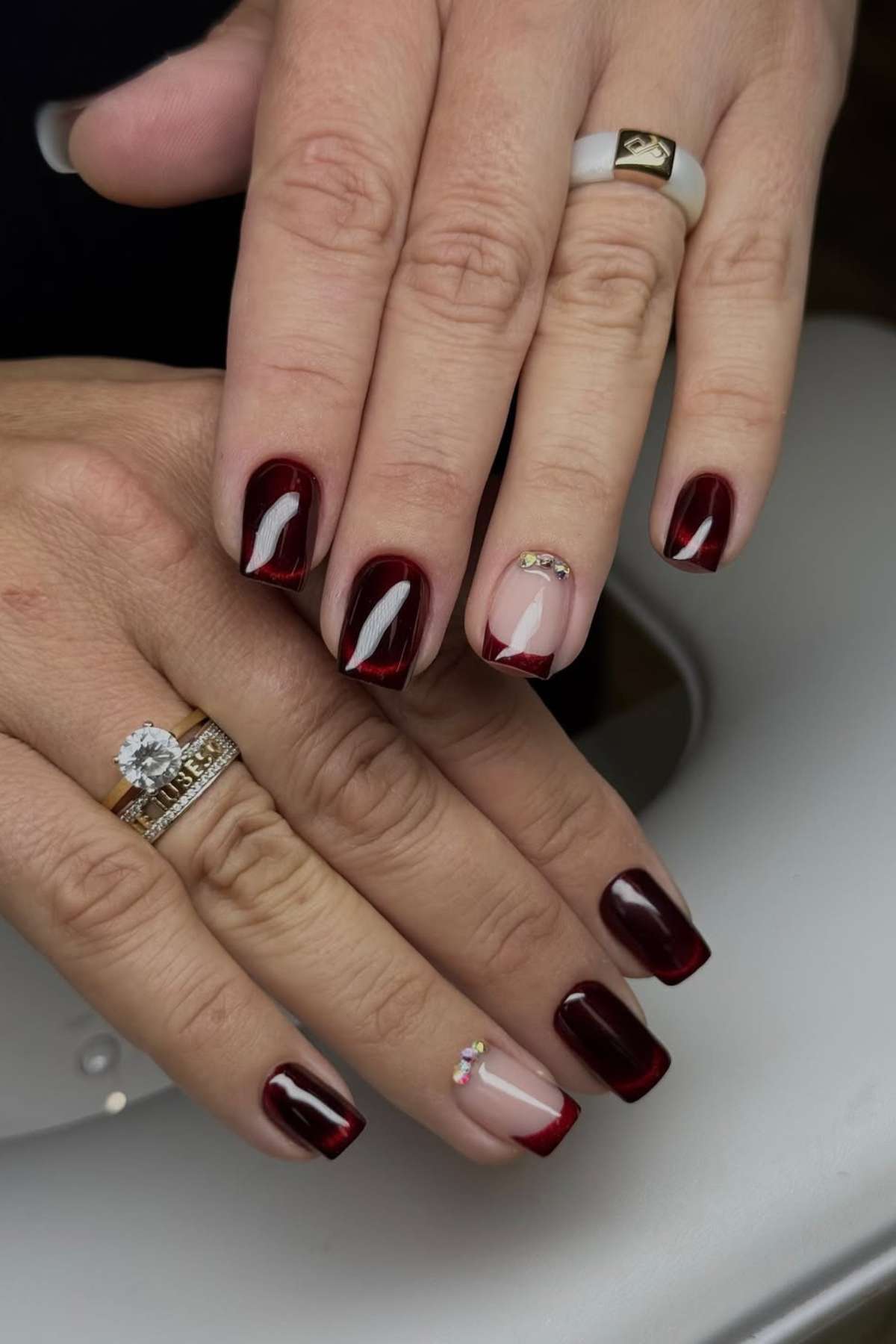Hands with manicured nails in dark red polish, featuring one accent nail on each hand with a clear base, red tip, and small rhinestone decorations. Rings are worn on both hands.