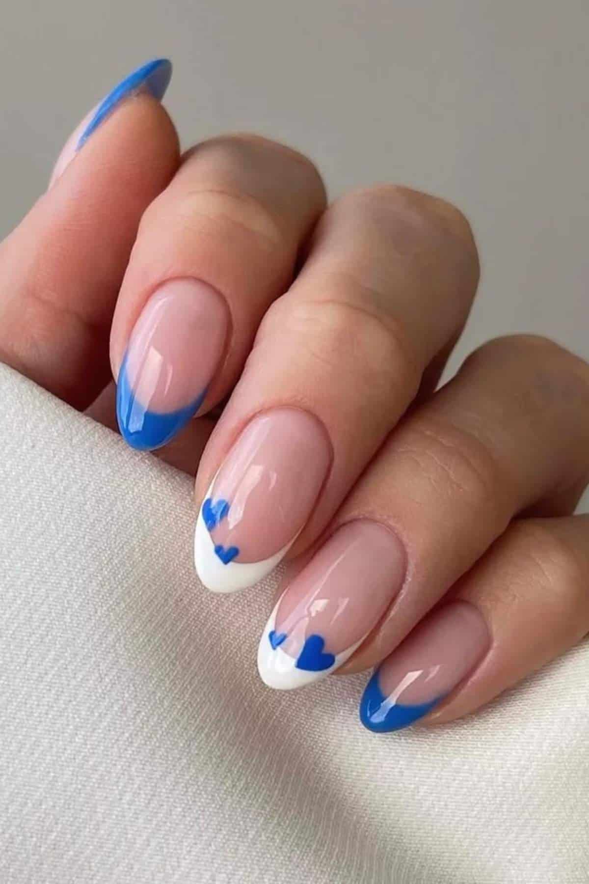 A hand with almond-shaped nails featuring blue French tips, white accents, and small blue heart designs on a light background.