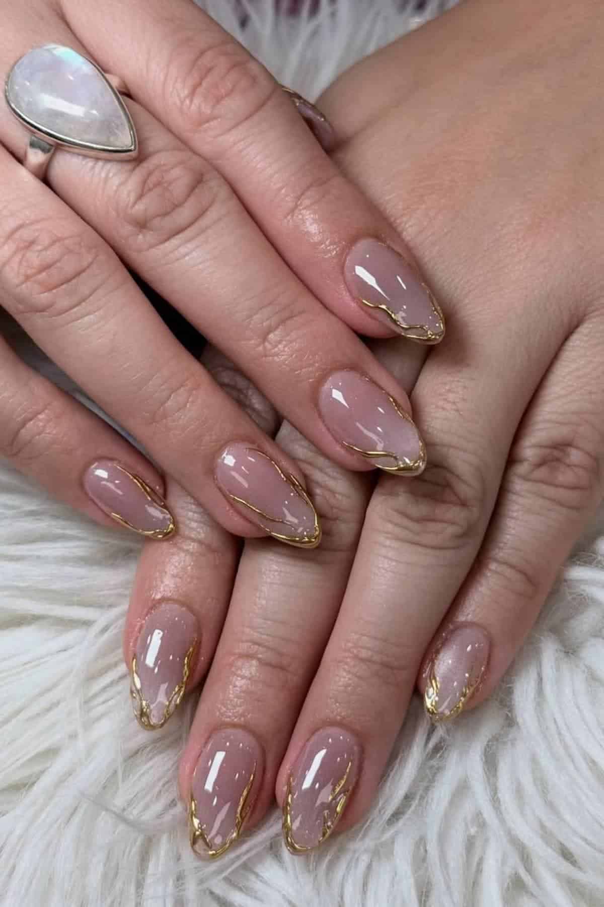 Hands with almond-shaped nails featuring a nude pink base and gold foil accents along the tips, displayed on a white furry surface. A silver ring with a teardrop-shaped stone is worn on one finger.