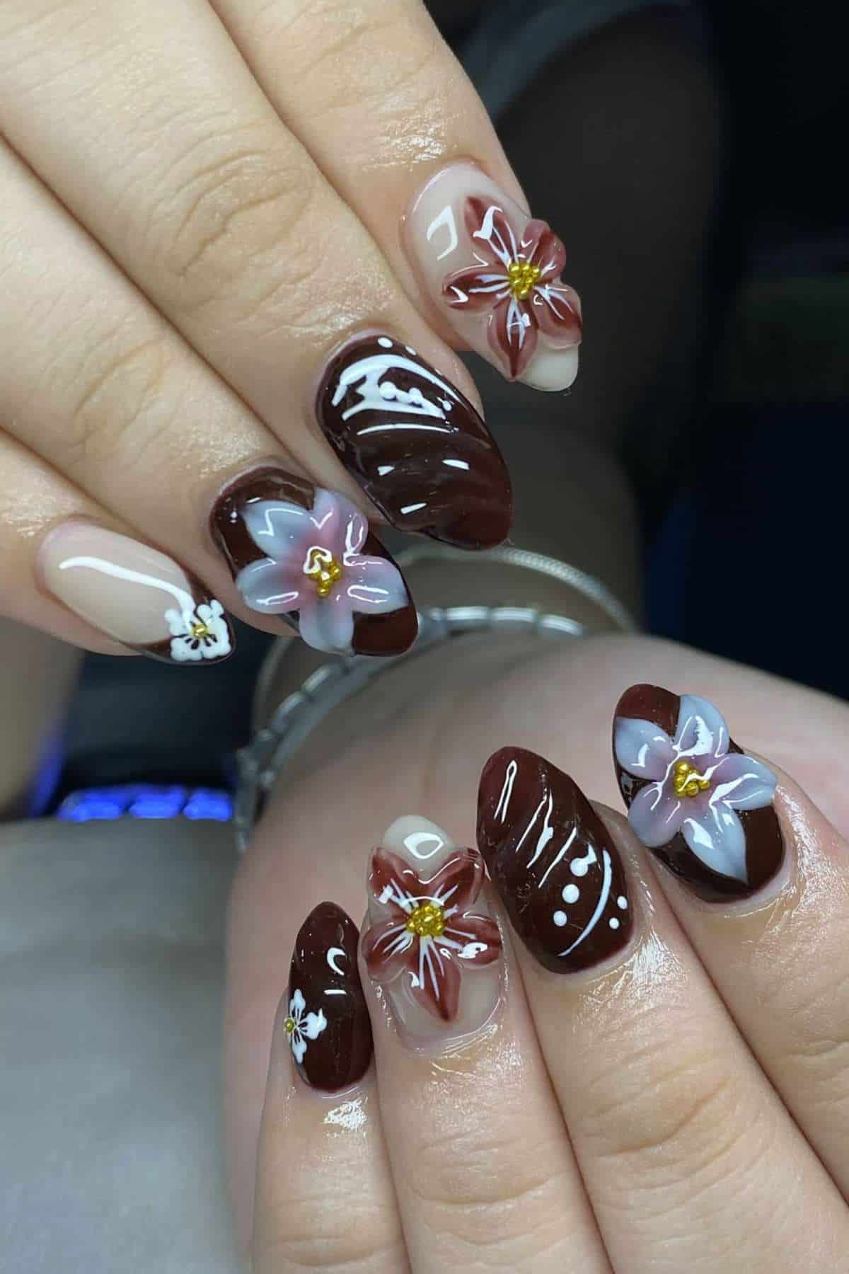 Hands with long, almond-shaped nails featuring glossy brown polish, white line accents, and 3D floral designs in pink and purple with yellow centers.
