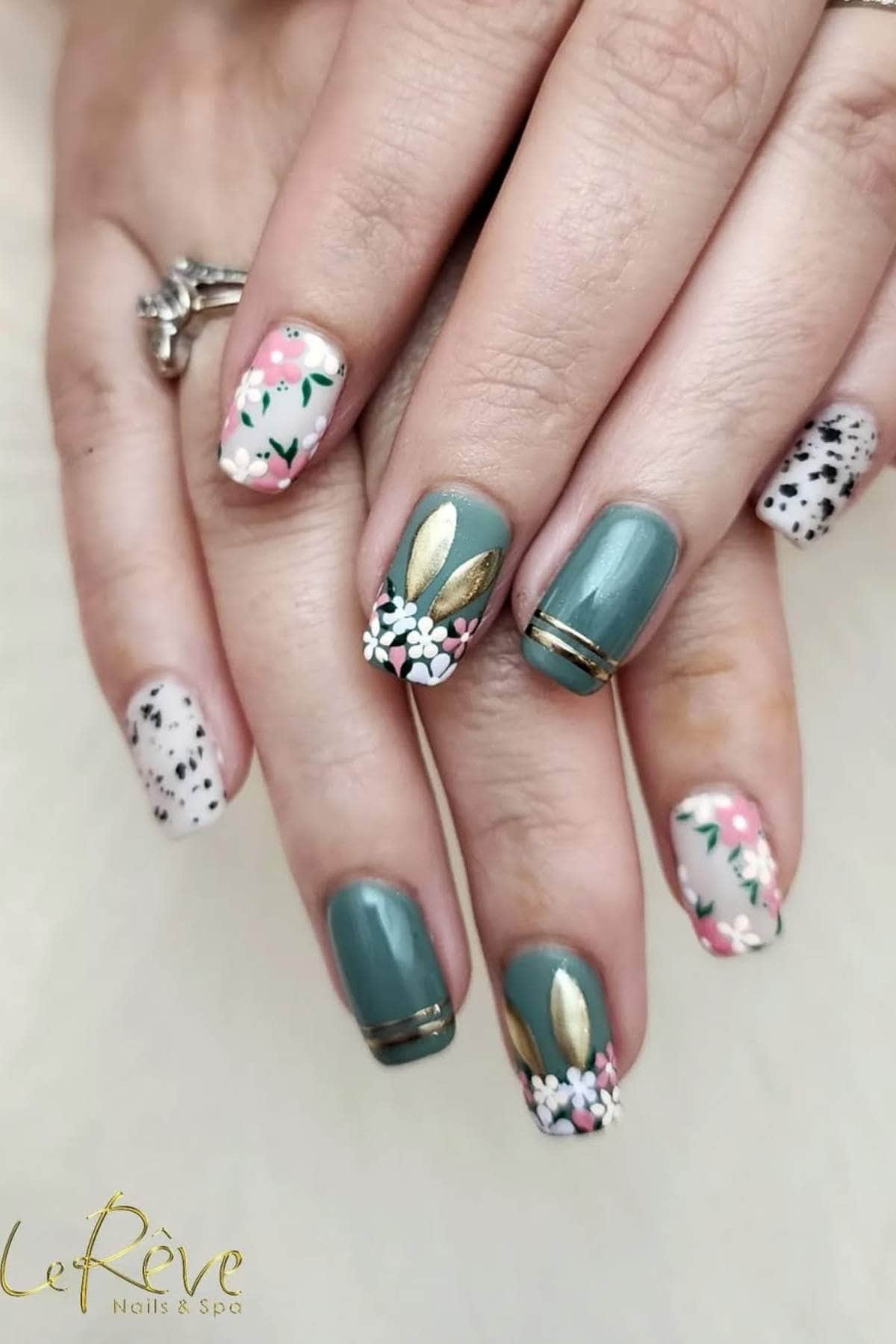 Hands with manicured nails featuring green polish, floral designs, gold leaf accents, and black-and-white speckled patterns. A ring is visible on one finger.