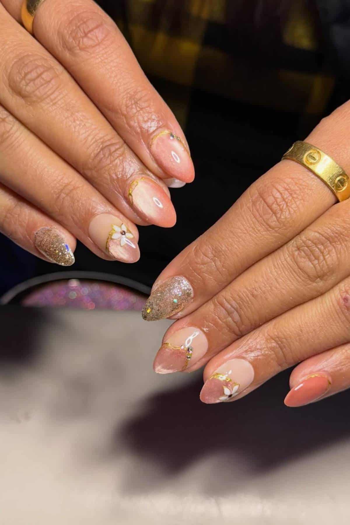 Hands with almond-shaped nails featuring a pink, gold glitter, and nude design, decorated with small white flowers and gold accents; wearer has gold rings on two fingers.